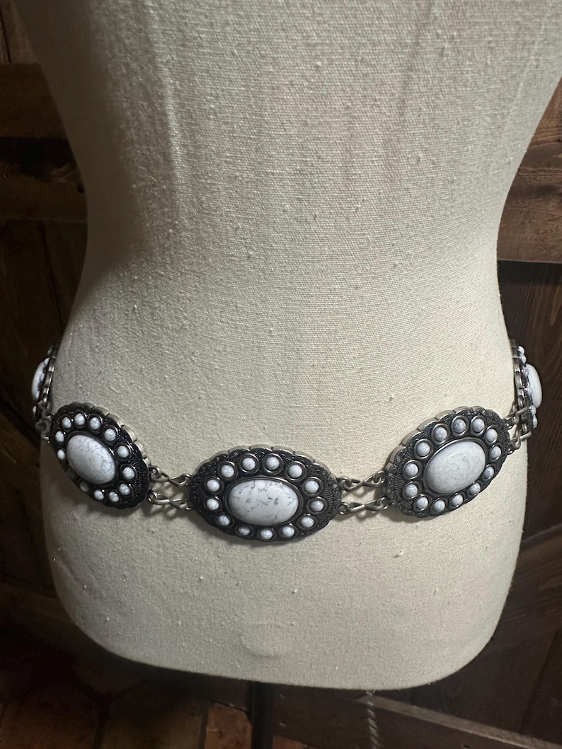 May include: A silver-toned belt with oval, white stone-like embellishments. Each oval is framed by a black border and surrounded by smaller white accents. The belt is displayed on a mannequin.