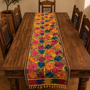 Puede incluir: Un vibrante camino de mesa bordado con un patrón floral repetido en tonos de rosa, naranja, azul y amarillo. El camino de mesa está colocado sobre una mesa de madera oscura, rodeada de sillas de madera. Los bordes tienen flecos. Mide unos 180 cm de largo.
