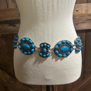 May include: A silver-toned belt with turquoise-colored oval and circular stones. The belt is displayed on a mannequin and has a western or bohemian style. The stones are set in a decorative metal frame.