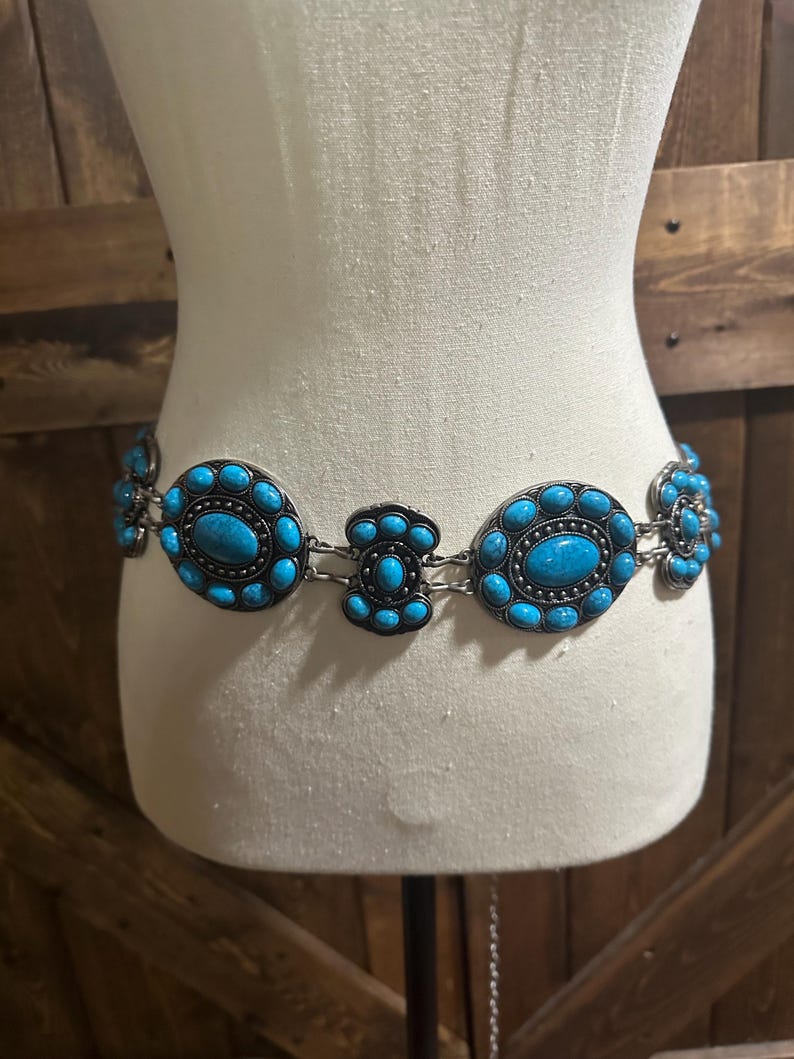 May include: A silver-toned belt with turquoise-colored oval and circular stones. The belt is displayed on a mannequin and has a western or bohemian style. The stones are set in a decorative metal frame.