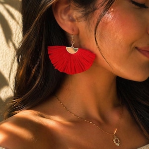 Puede incluir: Pendientes de borlas rojas con detalles dorados. Los pendientes tienen forma de abanico y cuelgan de pequeños aros dorados. También se ve un collar dorado con un colgante de sol.