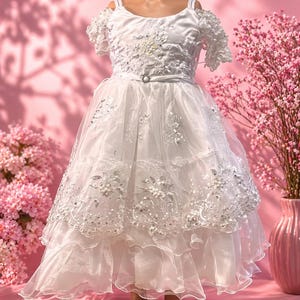 May include: A white formal dress with cold shoulder sleeves. The bodice is embellished with floral embroidery and a jeweled belt. The layered skirt features delicate lace and sequin details. The dress is displayed on a mannequin.