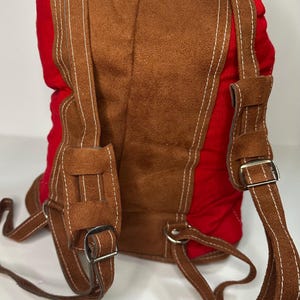 May include: Red and brown backpack with embroidered floral design. The backpack features brown suede-like material on the front and straps, with white stitching. The straps have metal buckles for adjustment. The back and sides are red.