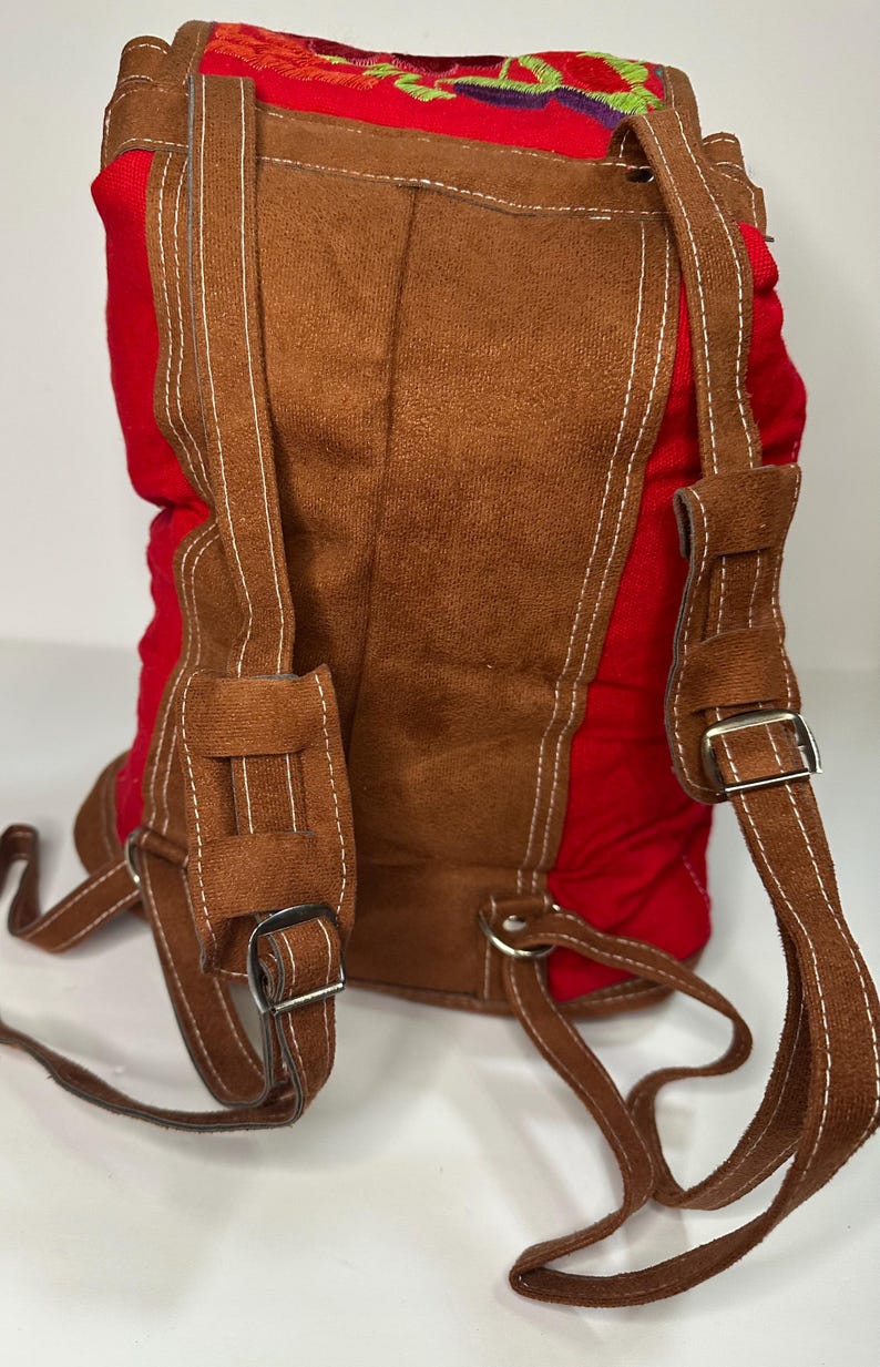 May include: Red and brown backpack with embroidered floral design. The backpack features brown suede-like material on the front and straps, with white stitching. The straps have metal buckles for adjustment. The back and sides are red.