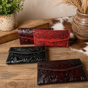 Puede incluir: Cuatro carteras de cuero en tonos rojos y negros, dispuestas sobre una superficie de madera. Cada cartera presenta un diseño floral en relieve y un cierre de solapa. Las carteras varían en color, desde burdeos oscuro hasta rojo brillante.