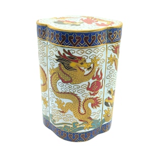Pode incluir: Um recipiente hexagonal em cloisonné com fundo branco e motivos de dragões em dourado, vermelho e preto. A tampa e a base são decoradas com uma borda azul profunda contendo padrões repetitivos em forma de coração em esmalte dourado e vermelho escuro.