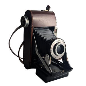 Op de afbeelding: Een vintage Kodak vouwcamera met een bruine lederen tas, een zwarte accordeonbalg en een ronde lensunit met zilverkleurige metalen accenten. Het apparaat is voorzien van een metalen steunframe en een lederen riem aan de zijkant.