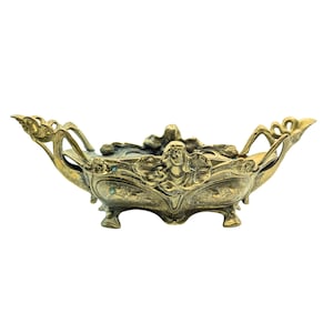 May include: An elongated brass bowl featuring an ornate relief design of a central female figure surrounded by flowing floral motifs. The vessel is supported by four small feet and includes two extended, curved handles on each side with a metallic finish.