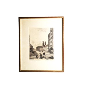Pode incluir: Uma gravura emoldurada a preto e branco de uma cena de rua de uma cidade europeia. A obra de arte retrata edifícios, pessoas e uma igreja com torres gémeas. A moldura é dourada e a obra de arte é mate.
