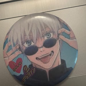 May include: Round button featuring an anime character with white hair and blue eyes wearing sunglasses. The button has the text "GOJO" inside a red heart and "INFINITY" on the bottom.