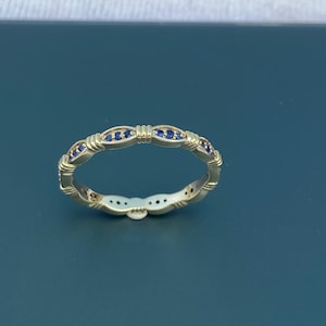 Peut inclure: Une bague dorée ornée d'un motif répétitif de pierres précieuses bleues. La bague présente un design délicat avec de petites pierres espacées. La bague est posée sur une surface bleu foncé.