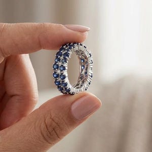 May include: A silver eternity band featuring two parallel rows of round, deep blue sapphire gemstones. The ring is constructed with a metallic setting that holds the stones in a continuous circular pattern, showcasing a polished finish throughout.