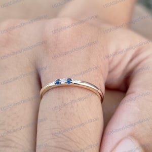 Puede incluir: Un anillo dorado con tres pequeñas piedras: una piedra blanca central flanqueada por dos piedras azules. El anillo está en un dedo, centrándose en el diseño y los detalles del anillo. La banda del anillo es lisa y pulida.