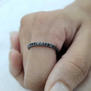 May include: A black ring with a band of small, black gemstones. The ring is worn on a finger, showcasing the dark stones against the skin. The ring's design is simple and elegant, suitable for everyday wear.