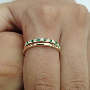 Op de afbeelding: Twee gouden ringen op een vinger gestapeld. De bovenste ring heeft een rij kleine, ronde, groene edelstenen afgewisseld met heldere edelstenen. De onderste ring is een eenvoudige gouden band.