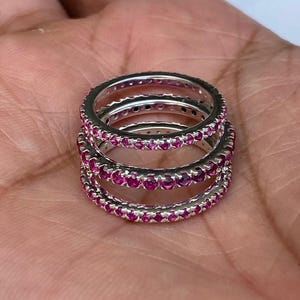 Può includere: Tre anelli d'argento impilati, ciascuno ornato da una fila continua di piccole e vibranti pietre preziose rosa. Gli anelli sono presentati su uno sfondo neutro, evidenziando il loro design e colore intricati.