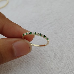 Pode incluir: Um anel de banda dourada com pedras preciosas verdes e brancas alternadas. O anel é segurado entre os dedos contra um fundo branco. As pedras são pequenas e espaçadas.