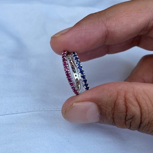 Könnte beinhalten: Zwei silberne Ringe, einer mit einer Reihe roter Edelsteine und der andere mit blauen Edelsteinen. Die Ringe werden zwischen den Fingern vor einem weißen Hintergrund gehalten. Die Edelsteine sind klein und eng gesetzt.