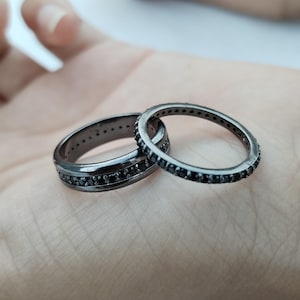 Può includere: Due anelli neri con piccole pietre nere incastonate attorno alle fasce. Un anello è più spesso dell'altro. Gli anelli sono appoggiati su una superficie chiara.