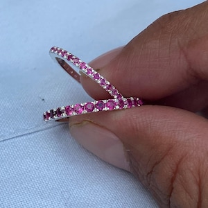 Może przedstawiać: Dwa srebrne pierścionki z ciągłym rzędem małych, jasnoróżowych kamieni szlachetnych. Pierścionki są trzymane na jasnoniebieskim tle. Kamienie szlachetne są równomiernie rozmieszczone i odbijają światło.