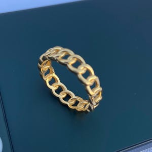 May include: A gold-colored ring with a chain link design. The ring is made of a shiny, metallic material and is displayed on a dark surface. The ring's links are evenly spaced, creating a continuous pattern.