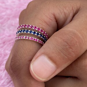 Può includere: Tre anelli d'argento con pietre preziose colorate. Due anelli presentano pietre rosa e un anello ha pietre blu. Gli anelli sono impilati su un dito, mostrando il design e le variazioni di colore dei gioielli.