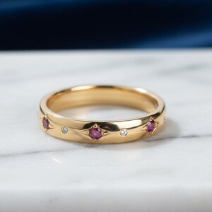 Könnte beinhalten: Ein goldener Bandring mit drei roten Edelsteinen und zwei kleinen, klaren Edelsteinen. Der Ring hat eine polierte Oberfläche und liegt auf einer weißen Marmoroberfläche.