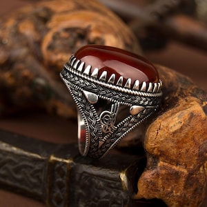 May include: A silver ring featuring a deep red, oval gemstone. The ring has an intricate, detailed design with a braided band and decorative features. The gemstone is set in a bezel with triangular accents.