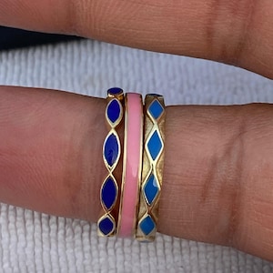 Op de afbeelding: Drie gouden ringen met kleurrijke emaille ontwerpen. Eén ring heeft een roze band, terwijl de andere blauw emaille hebben in diamant- en ovale vormen. De ringen zijn op een vinger gestapeld.