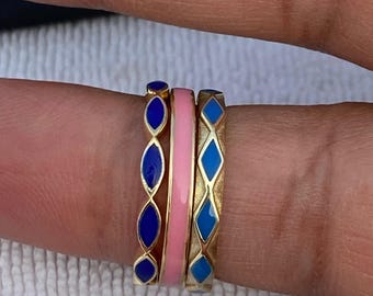 May include: Three gold rings with colourful enamel designs. One ring features a pink band, while the others have blue enamel in diamond and oval shapes. The rings are stacked on a finger.