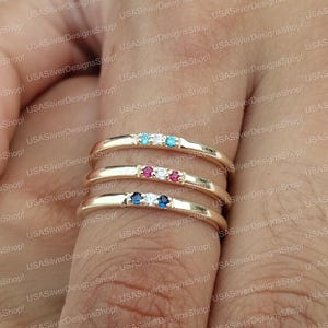 Puede incluir: Tres anillos dorados, cada uno con una fila de tres pequeñas piedras preciosas. El anillo superior tiene piedras turquesas y blancas, el anillo central tiene piedras rojas y blancas, y el anillo inferior tiene piedras azules y blancas.