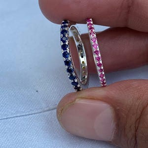 Può includere: Vengono mostrati tre anelli d'argento, ciascuno con un diverso colore di pietra preziosa. Un anello presenta gemme blu, un altro ha gemme rosa e il terzo gemme trasparenti. Gli anelli sono tenuti dalle dita di una persona.