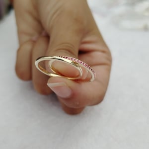 Peut inclure: Deux bagues en or, dont une avec une rangée de petites pierres précieuses rouges. Les bagues sont entrelacées, créant un design unique et élégant. Les bagues sont tenues dans une main, mettant en valeur leurs détails délicats.