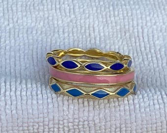 May include: Three gold-coloured rings stacked together. The top ring has a scalloped edge with blue enamel details. The middle ring is pink enamel. The bottom ring has a geometric design with blue enamel.