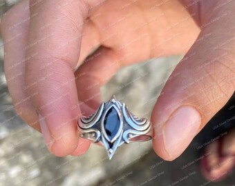 May include: A silver ring with an ornate design, featuring a central black gemstone. The ring has a detailed, symmetrical pattern with curved lines and pointed elements. The ring is held in a hand, showcasing its intricate craftsmanship.