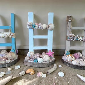 May include: Three decorative wooden ladders painted in shades of blue and gray, adorned with seashells, starfish, and pearls. The ladders are set on a base of wood and shells, creating a coastal theme. Each ladder has a unique design.