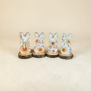 May include: Four wooden bunny figurines with a sunflower design. Each bunny is mounted on a small, round wooden base. The bunnies have a light blue background with yellow sunflowers and are decorated with twine bows.
