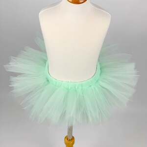 May include: A light green tulle tutu skirt on a white mannequin. The skirt is gathered at the waist and has a soft, flowing look.