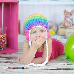 May include: A young girl wearing a rainbow crochet hat with white crocheted ties, covering her mouth with her hand, lying on a white wooden surface with colorful candy scattered around her.