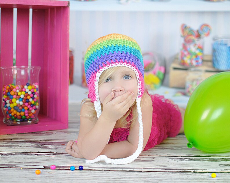 May include: A young girl wearing a rainbow crochet hat with white crocheted ties, covering her mouth with her hand, lying on a white wooden surface with colorful candy scattered around her.