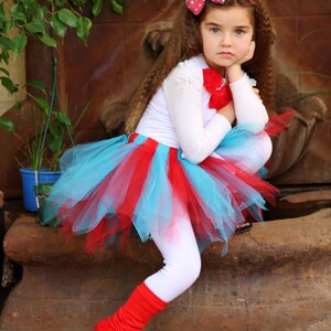 May include: A young girl wearing a white long-sleeved shirt, a red and blue tulle tutu skirt, red leg warmers, and black shoes. She is sitting on a stone surface with her hand on her chin and looking down.