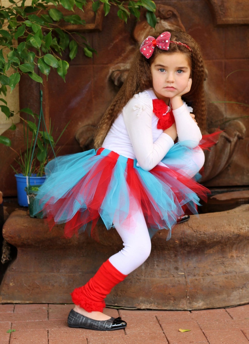 May include: A young girl wearing a white long-sleeved shirt, a red and blue tulle tutu skirt, red leg warmers, and black shoes. She is sitting on a stone surface with her hand on her chin and looking down.