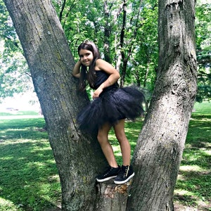 May include: A young girl wearing a black tulle tutu skirt and a black tank top, standing on a tree trunk in a park.