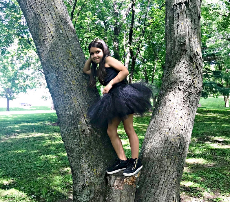 May include: A young girl wearing a black tulle tutu skirt and a black tank top, standing on a tree trunk in a park.