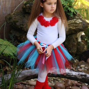 May include: A young girl wearing a white shirt with a red flower collar, a red and blue tulle tutu skirt, red leg warmers, and a pink polka dot bow in her hair.