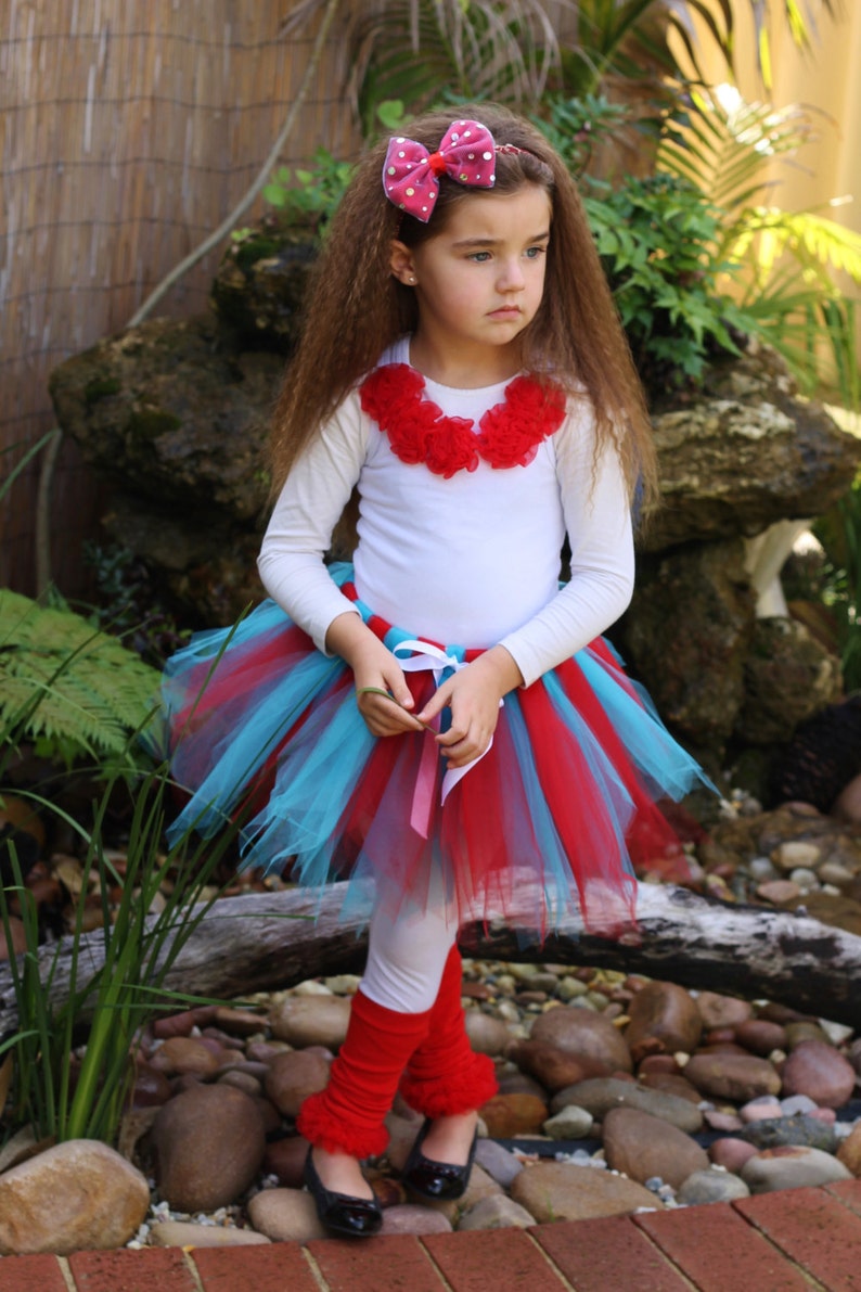 May include: A young girl wearing a white shirt with a red flower collar, a red and blue tulle tutu skirt, red leg warmers, and a pink polka dot bow in her hair.