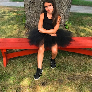 May include: A young girl wearing a black tank top and a black tulle tutu skirt sits on a red bench. She is wearing a gold cat ear headband with pearl accents. The girl is sitting on a grassy lawn with a large tree behind her.