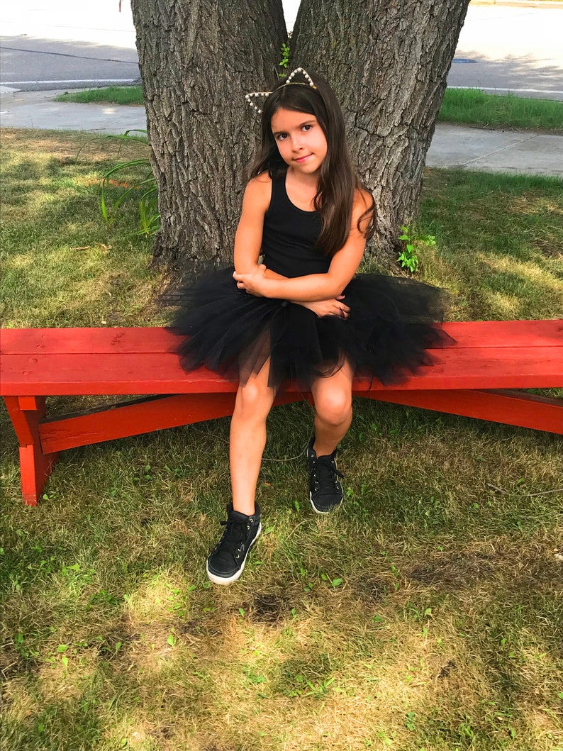 May include: A young girl wearing a black tank top and a black tulle tutu skirt sits on a red bench. She is wearing a gold cat ear headband with pearl accents. The girl is sitting on a grassy lawn with a large tree behind her.