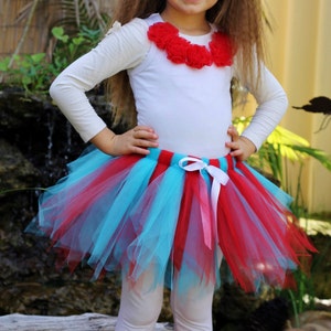 May include: A young girl wearing a white long-sleeved shirt with a red flower embellishment, a turquoise and red tulle tutu skirt, white leggings, and red leg warmers with a fuzzy trim. She is wearing a pink and white polka dot bow headband.