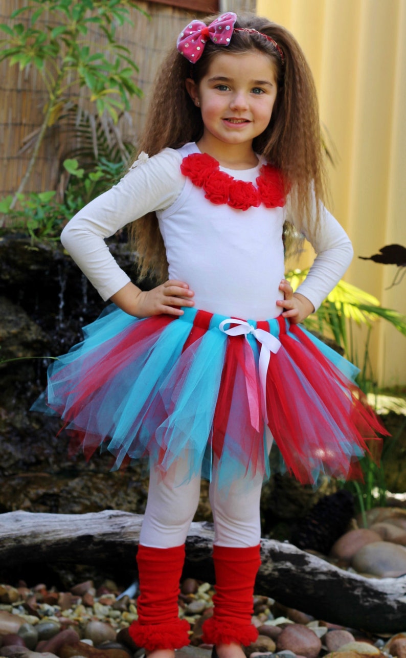 May include: A young girl wearing a white long-sleeved shirt with a red flower embellishment, a turquoise and red tulle tutu skirt, white leggings, and red leg warmers with a fuzzy trim. She is wearing a pink and white polka dot bow headband.